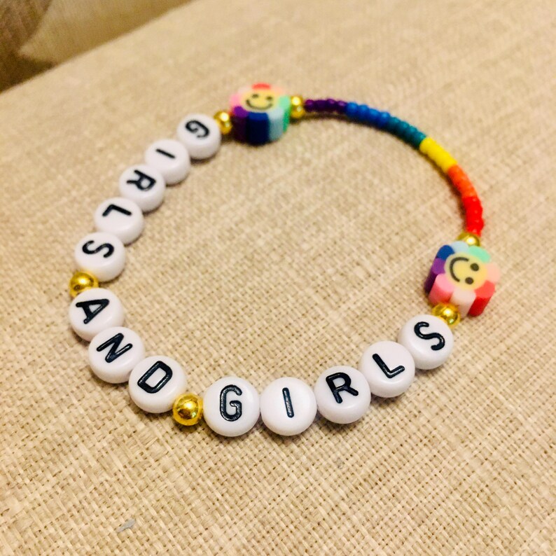 Op de afbeelding: Een kralenarmband met witte ronde kralen met zwarte letters die GIRLS AND GIRLS spellen, afgewisseld met kleine goudkleurige metalen kralen, twee veelkleurige bloemvormige kralen met smileygezichtjes en een segment van regenboogkleurige kraaltjes.