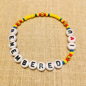 May include: A circular beaded bracelet featuring white alphabet beads spelling out REMEMBERED and UAI with a red heart bead. The design includes small yellow, red, and blue seed beads interspersed with metallic gold spherical beads throughout the strand.