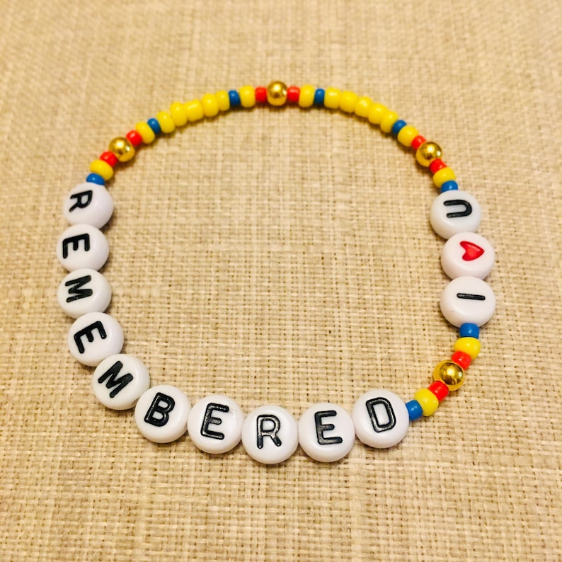 May include: A circular beaded bracelet featuring white alphabet beads spelling out REMEMBERED and UAI with a red heart bead. The design includes small yellow, red, and blue seed beads interspersed with metallic gold spherical beads throughout the strand.