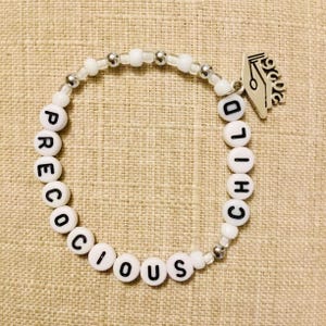 May include: White beaded bracelet spelling "PRECIOUS CHILD" with silver accents and a silver pen charm. The bracelet is made of white beads and silver-coloured beads. The bracelet is on a tan fabric background.