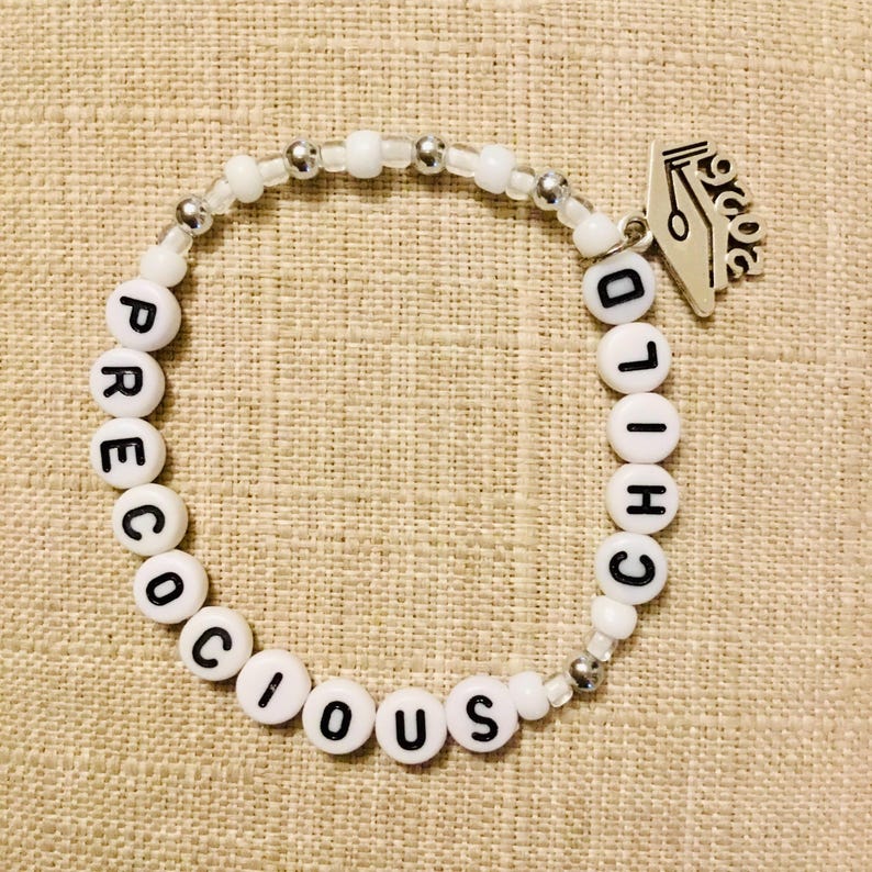 May include: White beaded bracelet spelling "PRECIOUS CHILD" with silver accents and a silver pen charm. The bracelet is made of white beads and silver-coloured beads. The bracelet is on a tan fabric background.