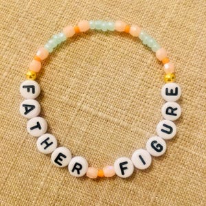 May include: A beaded bracelet with the words "FATHER FIGURE" spelled out in black letters on white round beads. The bracelet features a mix of peach, orange, and light green beads, with gold-colored accents.