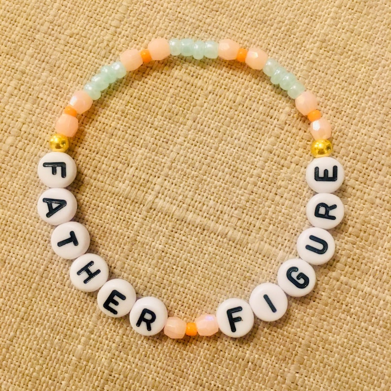 May include: A beaded bracelet with the words "FATHER FIGURE" spelled out in black letters on white round beads. The bracelet features a mix of peach, orange, and light green beads, with gold-colored accents.