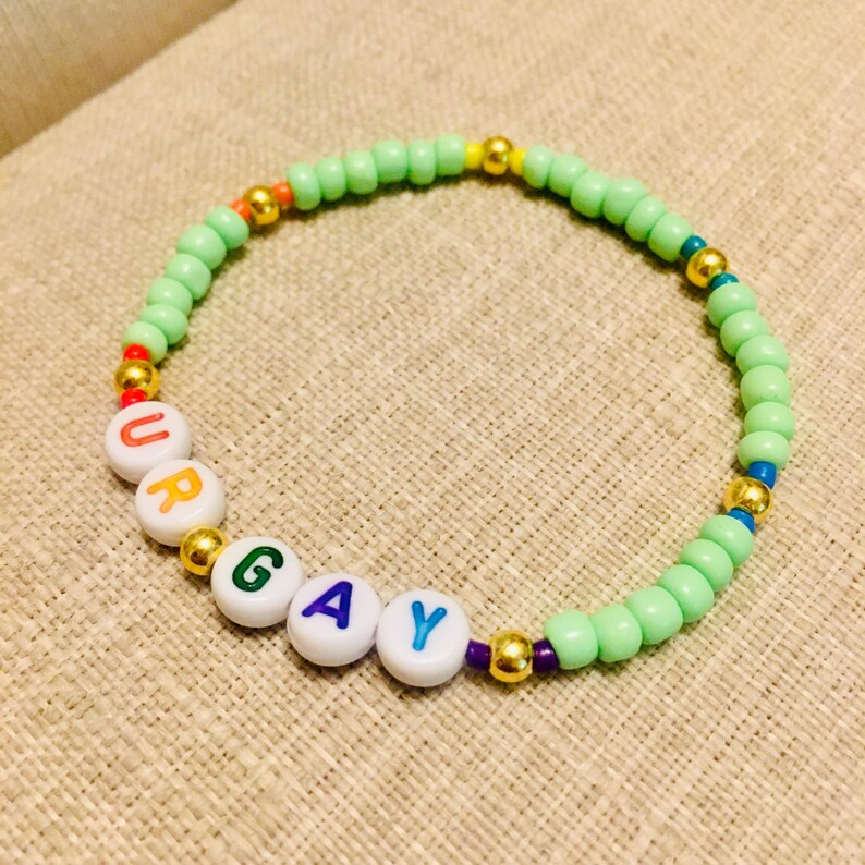 Op de afbeelding: Een kralenarmband met lichtgroene ronde kralen afgewisseld met kleine metallic gouden bolletjes. De armband bevat witte ronde kralen met gekleurde letters die het woord URGAY spellen, waarbij elke letter een andere kleur heeft.