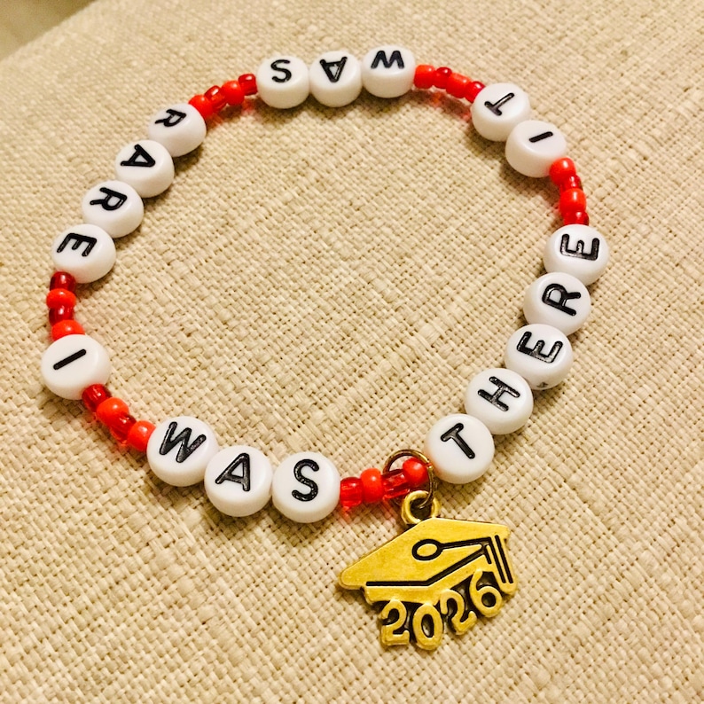 May include: A beaded bracelet with white letter beads spelling "I WAS THERE" and a gold graduation cap charm with "2026". The bracelet features red beads and is on a beige surface.