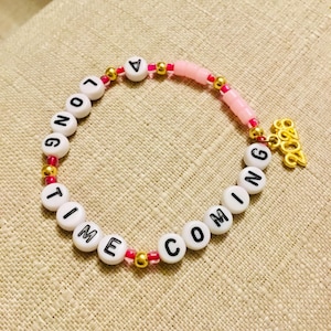 May include: A bracelet made of white letter beads spelling "LONG TIME COMING". The bracelet has pink and gold beads, and a gold charm with the number "2022".