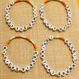 May include: Four beaded bracelets with white letter beads spelling out phrases. The bracelets have orange, green, and gold accents. The phrases include "PROTECT THE FAMILY", "STEP INTO MY OFFICE", "DICKS BIGGER", and "DRINK THAT BROWN LIQUOR".