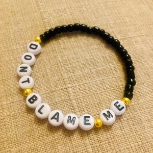 May include: A beaded bracelet with the words "DON'T BLAME ME" spelled out in black letters on white beads. The bracelet also features black beads and gold-colored accent beads. The bracelet is displayed on a tan surface.