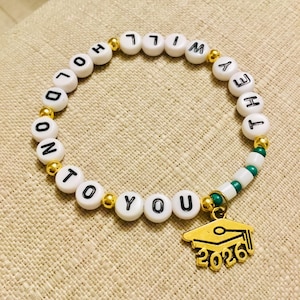 Puede incluir: Pulsera de cuentas blancas con cuentas de letras negras que deletrean "HOLD ON TO YOU". Tambi&eacute;n presenta detalles dorados y un dije de gorro de graduaci&oacute;n dorado con "2026". La pulsera est&aacute; sobre una superficie beige.
