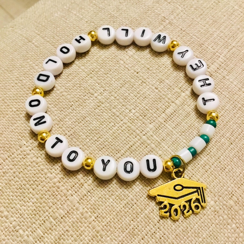 Puede incluir: Pulsera de cuentas blancas con cuentas de letras negras que deletrean "HOLD ON TO YOU". Tambi&eacute;n presenta detalles dorados y un dije de gorro de graduaci&oacute;n dorado con "2026". La pulsera est&aacute; sobre una superficie beige.