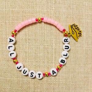 May include: A pink beaded bracelet with the words "ALL JUST A BLUR" spelled out in white letter beads. The bracelet has gold and pink accents and a gold-coloured pen nib charm.