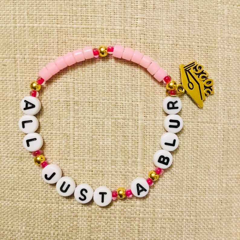 May include: A pink beaded bracelet with the words "ALL JUST A BLUR" spelled out in white letter beads. The bracelet has gold and pink accents and a gold-coloured pen nib charm.