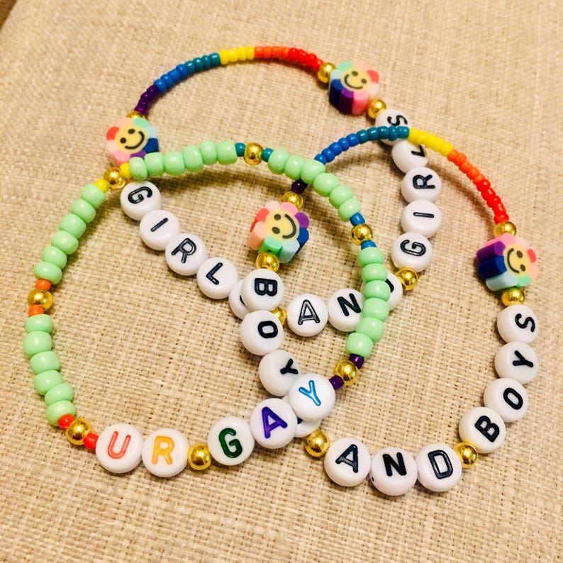 Op de afbeelding: Twee kralenarmbanden met witte letterkralen die "GIRL AND BOYS" en "UR GAY AND BOYS" spellen. De armbanden bevatten lichtgroene, regenboogkleurige en goudkleurige kralen, samen met veelkleurige bloemvormige kralen met lachende gezichtjes.