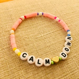 Op de afbeelding: Een kralenarmband met een reeks lichtroze cilindrische kralen afgewisseld met kleine witte, gele, oranje en blauwe accentkralen. De armband bevat witte letterkralen die CALM DOWN spellen en een kleine goudkleurige hartvormige kraal.