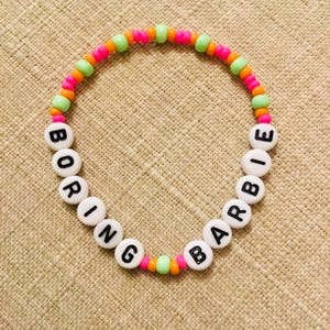 May include: A beaded bracelet with the words "BORING BARBIE" spelled out in black letters on white beads. The bracelet features a colorful design with pink, orange, and green beads. The bracelet is on a tan surface.