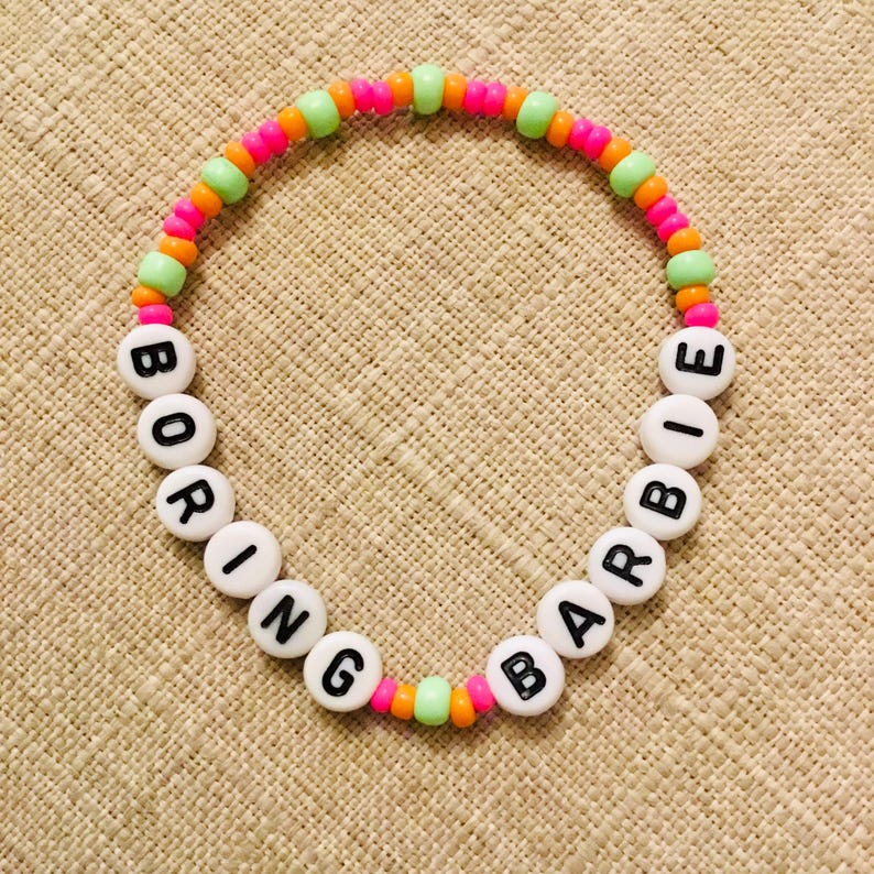 May include: A beaded bracelet with the words "BORING BARBIE" spelled out in black letters on white beads. The bracelet features a colorful design with pink, orange, and green beads. The bracelet is on a tan surface.