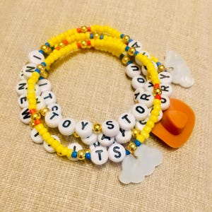 Op de afbeelding: Een set kralenarmbanden met gele, rode en blauwe rocailles afgewisseld met goudkleurige tussenkralen. Witte letterkralen vormen de tekst TOY STORY. De armbanden bevatten een bruine cowboyhoed-bedel en twee witte, wolk-vormige bedels.