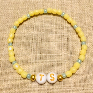 May include: A circular beaded bracelet featuring alternating yellow and light blue beads. Two white spherical beads with the orange letters T and S are positioned at the bottom, flanked by two small metallic gold beads on either side of the letters.