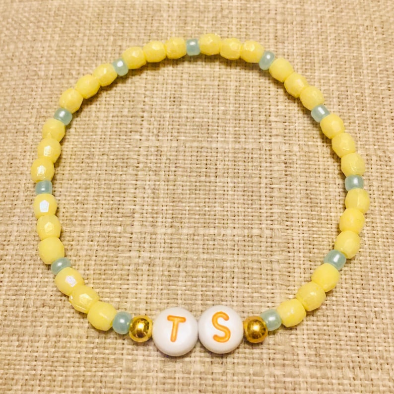 May include: A circular beaded bracelet featuring alternating yellow and light blue beads. Two white spherical beads with the orange letters T and S are positioned at the bottom, flanked by two small metallic gold beads on either side of the letters.