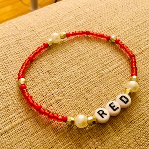 Puede incluir: Una pulsera de cuentas compuesta por peque&ntilde;as cuentas de vidrio rojas, intercaladas con perlas blancas y cuentas espaciadoras met&aacute;licas doradas. Incluye tres cuentas circulares blancas con las letras R, E y D impresas en negro formando RED.