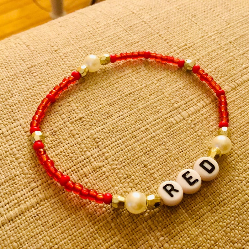 Puede incluir: Una pulsera de cuentas compuesta por peque&ntilde;as cuentas de vidrio rojas, intercaladas con perlas blancas y cuentas espaciadoras met&aacute;licas doradas. Incluye tres cuentas circulares blancas con las letras R, E y D impresas en negro formando RED.