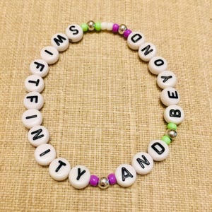 May include: A circular beaded bracelet featuring white round beads with black letters spelling out SWIFTFINITY AND BEYOND. The design includes small lime green, purple, and metallic silver spacer beads interspersed between the lettered beads.