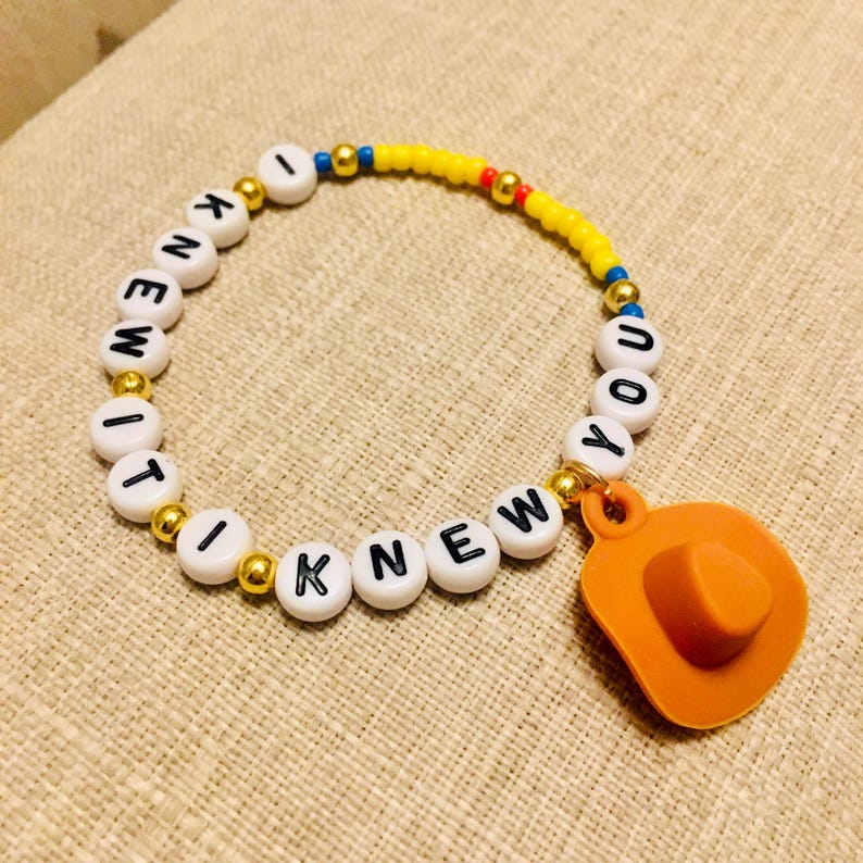 May include: A beaded bracelet featuring white round beads with black letters spelling out I KNEW IT I KNEW YOU, interspersed with small metallic gold beads, a section of yellow beads, and a single orange cowboy hat charm attached to the band.