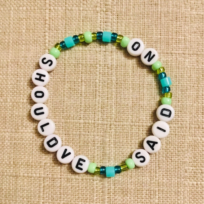 May include: A beaded bracelet with the words "SHOULD LOVE SAID ON" spelled out in white letter beads. The bracelet features turquoise, green, and clear beads interspersed between the letter beads. The bracelet is on a tan fabric surface.