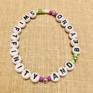 May include: A circular beaded bracelet featuring white round beads with black letters spelling out SWIFTFINITY AND BEYOND. The design includes small lime green, purple, and metallic silver spacer beads interspersed between the lettered beads.