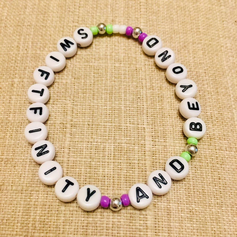 May include: A circular beaded bracelet featuring white round beads with black letters spelling out SWIFTFINITY AND BEYOND. The design includes small lime green, purple, and metallic silver spacer beads interspersed between the lettered beads.