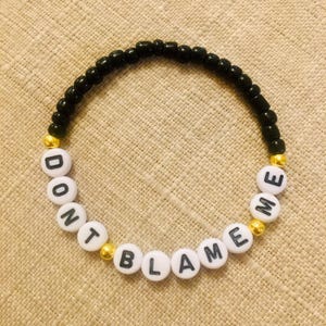 May include: A beaded bracelet with the words "DON'T BLAME ME" spelled out in white letter beads. The bracelet features black beads and gold-colored accent beads, creating a simple yet eye-catching design.