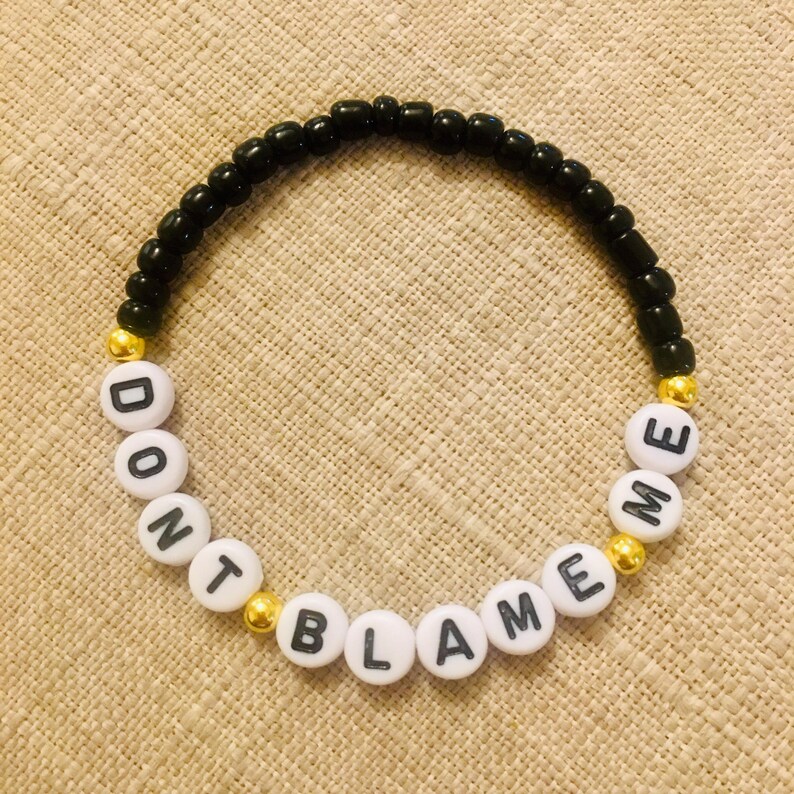 May include: A beaded bracelet with the words "DON'T BLAME ME" spelled out in white letter beads. The bracelet features black beads and gold-colored accent beads, creating a simple yet eye-catching design.