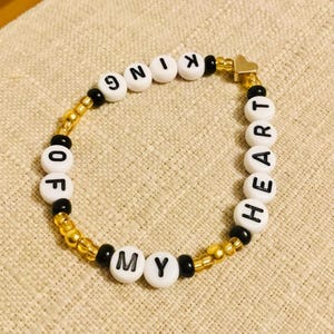 May include: A beaded bracelet with the words "KING OF MY HEART" spelled out in white letter beads. The bracelet features gold and black beads, and a small gold heart charm.