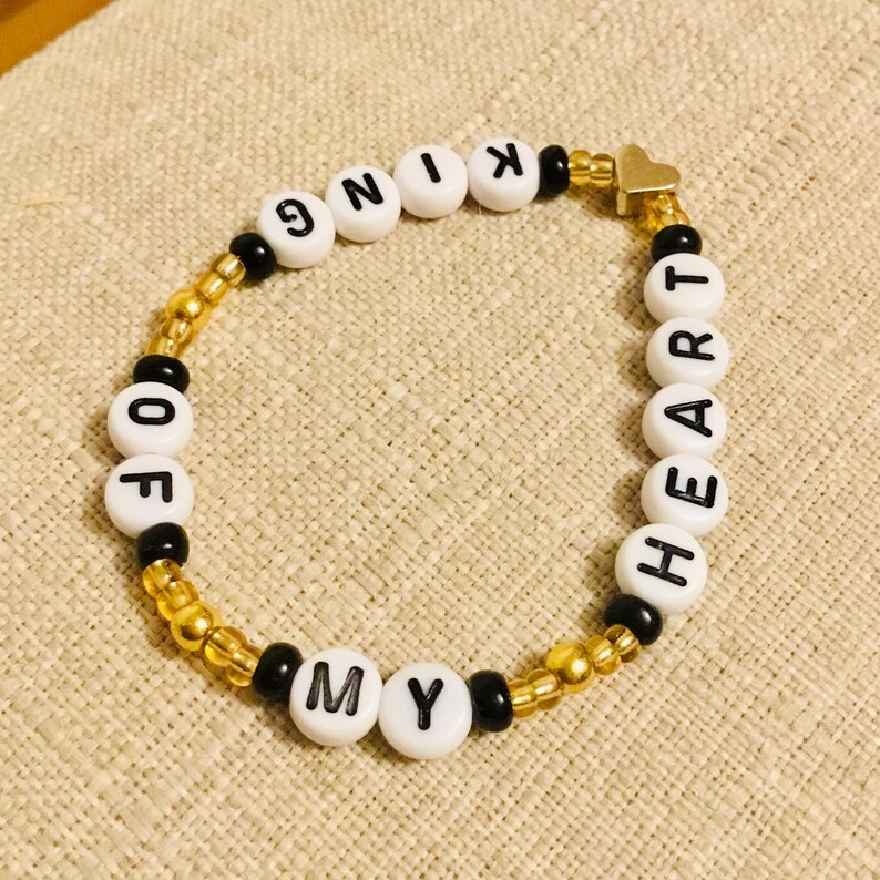 May include: A beaded bracelet with the words "KING OF MY HEART" spelled out in white letter beads. The bracelet features gold and black beads, and a small gold heart charm.