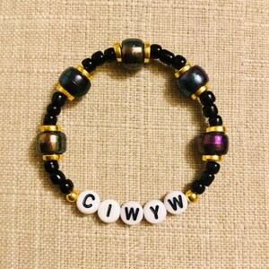 May include: A beaded bracelet with black, gold, and iridescent beads. The bracelet features the letters "CIWYW" on white beads. The iridescent beads have purple, green, and gold hues. The bracelet is on a tan surface.