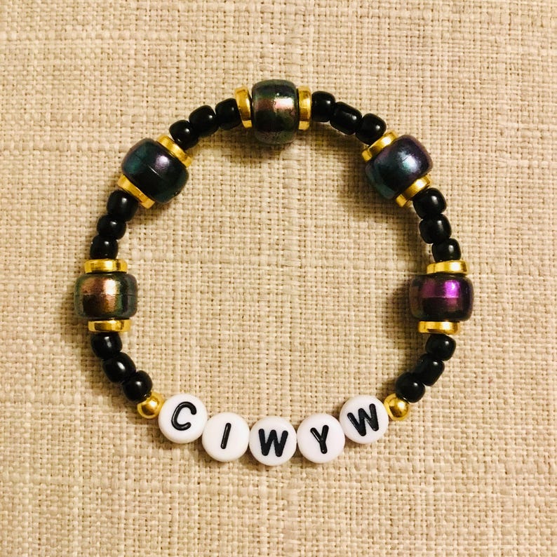 May include: A beaded bracelet with black, gold, and iridescent beads. The bracelet features the letters "CIWYW" on white beads. The iridescent beads have purple, green, and gold hues. The bracelet is on a tan surface.