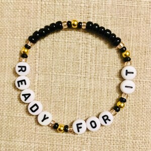 May include: A beaded bracelet with the words "READY FOR IT" spelled out in white letter beads. The bracelet features black, gold, and clear beads. The bracelet is displayed on a neutral-colored surface.