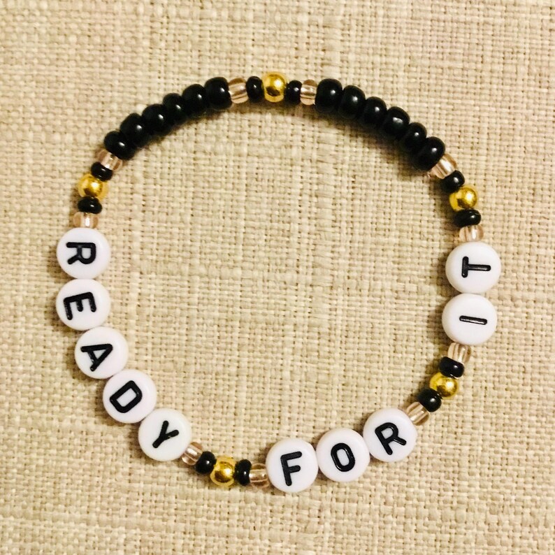 May include: A beaded bracelet with the words "READY FOR IT" spelled out in white letter beads. The bracelet features black, gold, and clear beads. The bracelet is displayed on a neutral-colored surface.