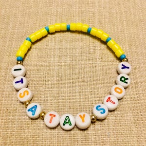 May include: A circular beaded bracelet featuring a sequence of bright yellow cylindrical beads interspersed with small blue beads. The lower section consists of white round beads with colorful letters spelling out the phrase ITS A TAY STORY.