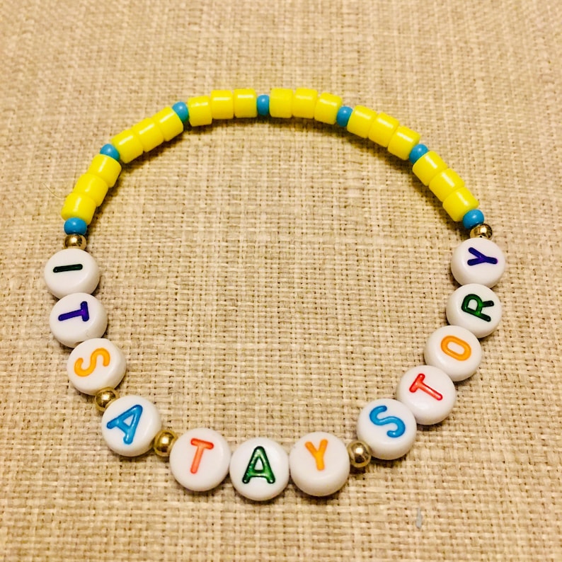 May include: A circular beaded bracelet featuring a sequence of bright yellow cylindrical beads interspersed with small blue beads. The lower section consists of white round beads with colorful letters spelling out the phrase ITS A TAY STORY.