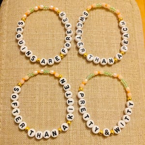 May include: Four beaded bracelets with white letter beads spelling out phrases like "Sequins are forever" and "Sweeter than peach." The bracelets have gold and pastel-colored accent beads.