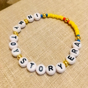 May include: A beaded bracelet featuring white round beads with black letters spelling out IN MY TOY STORY ERA, interspersed with metallic gold spherical beads, and a section of small yellow seed beads with a single red bead and blue accents.