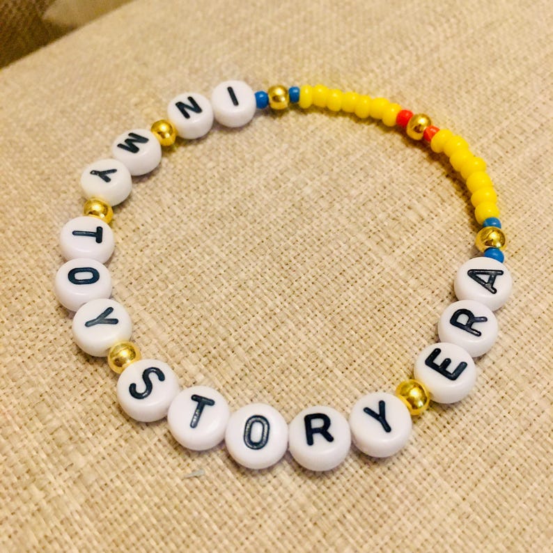 May include: A beaded bracelet featuring white round beads with black letters spelling out IN MY TOY STORY ERA, interspersed with metallic gold spherical beads, and a section of small yellow seed beads with a single red bead and blue accents.