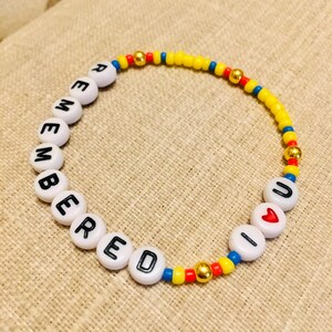 May include: A circular beaded bracelet featuring white letter beads spelling out REMEMBERED U, a red heart bead, small yellow, blue, and red seed beads, and metallic gold-toned spacer beads arranged in a repeating pattern along the elastic cord.
