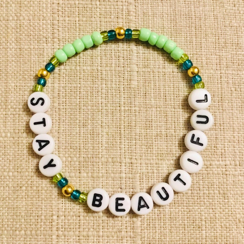 May include: A beaded bracelet with the words "STAY BEAUTIFUL" spelled out in white letter beads. The bracelet features green, gold, and teal beads. The bracelet is on a beige textured surface.