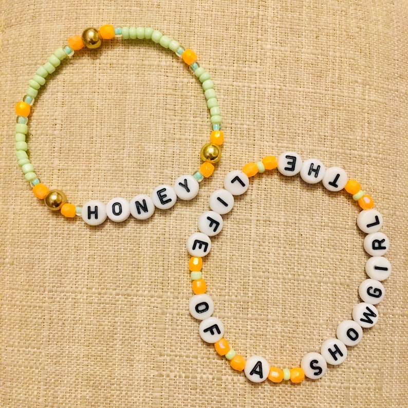 May include: Two beaded bracelets. One is green with the word "HONEY" spelled out in white letter beads. The other is orange with the words "A SHOW GIRL OF THE LIFE" spelled out in white letter beads. Gold and orange beads accent the bracelets.