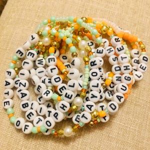 May include: A collection of beaded bracelets with white letter beads spelling out words like "FATE", "TAYLOR", and "DAUGHTER". The bracelets feature a mix of colors including orange, green, and gold, with pearl accents.