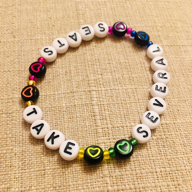 Op de afbeelding: Een cirkelvormige kralenarmband met witte ronde kralen met zwarte letters die de tekst TAKE SEVERAL SEATS vormen. Het ontwerp bevat zwarte kralen met kleurrijke hartomlijningen in roze, oranje, geel, groen en blauw, afgewisseld met kleine metallic en doorschijnende kralen.