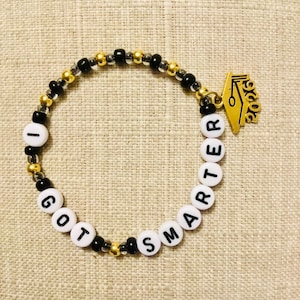 May include: A beaded bracelet with the words "I GOT SMARTER" spelled out in white letter beads. The bracelet features gold and black beads, and a gold graduation cap charm with "2026" on it.