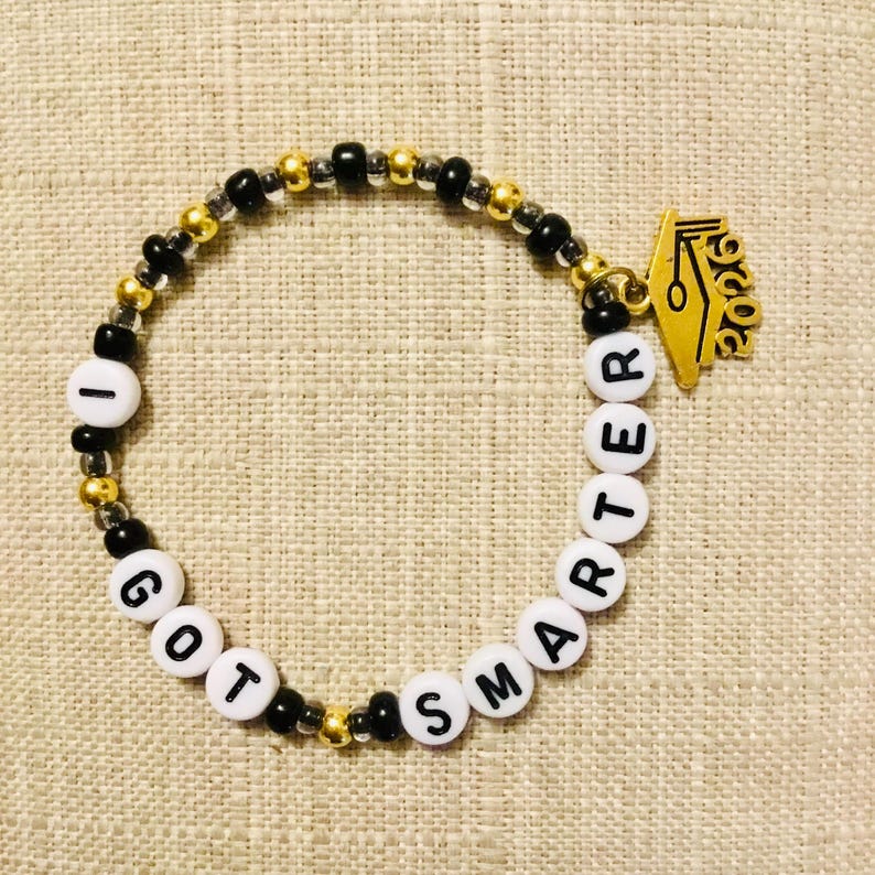 May include: A beaded bracelet with the words "I GOT SMARTER" spelled out in white letter beads. The bracelet features gold and black beads, and a gold graduation cap charm with "2026" on it.
